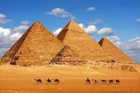 Camel caravan traveling across desert sand toward the pyramids of Giza under blue sky with white clouds